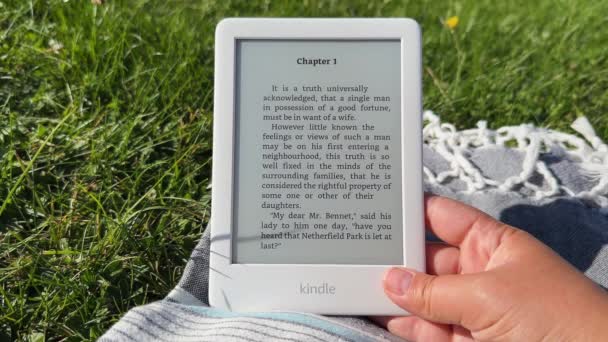 白色的亚马逊Kindle电子书阅读器手牵着手在户外草坪上的野餐毯上 在公园里看小说 波兰华沙 2023年9月15日 — 图库视频影像