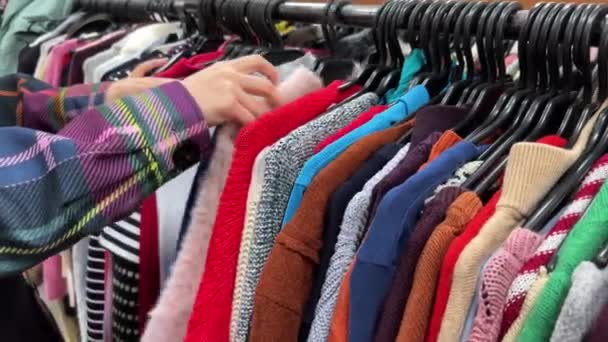若い女性は中古の店で服を選ぶ カラフルなセーターが付いている漂流店のハンガー 使用済みの服を購入するというコンセプト ファストファッション業界の廃棄物は減少し 持続可能性を高めます クローズアップ — ストック動画