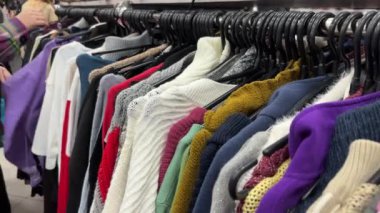 Genç bir kadın ikinci el dükkanından kıyafet alıyor. Renkli kazaklı ikinci el mağazası askıları. Hızlı moda endüstrisi israfı ve sürdürülebilirliği azaltır. Önplanda seçmeli odak.
