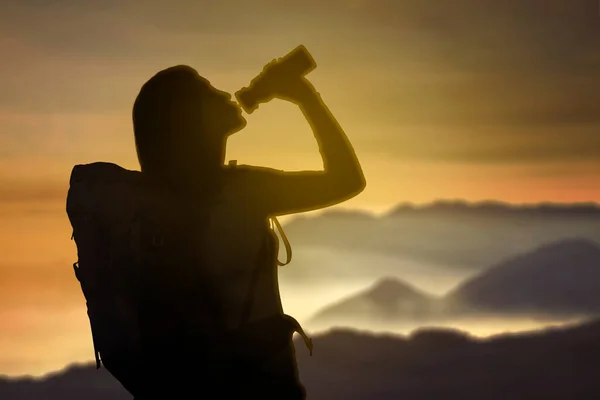 夜明けの日差しを背景に立ちながら山をハイキングした後に水を飲む女性ハイカーのシルエット — ストック写真