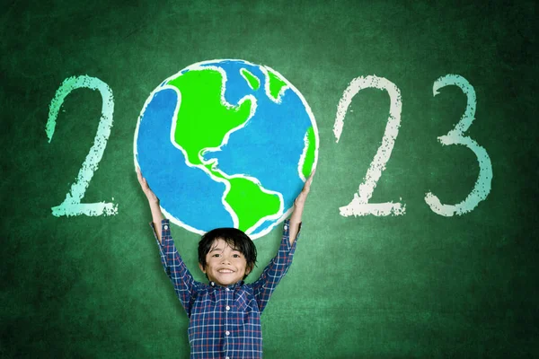 教室に立っている間に黒板に2023番で描かれた地球を持ち上げる幸せな男の子 — ストック写真