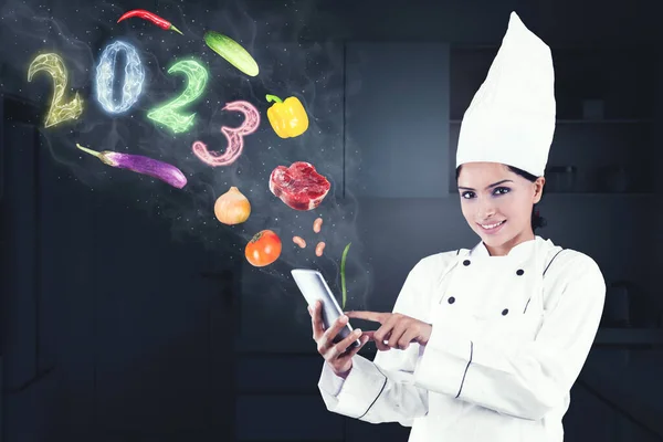 Image of female magician chef using a cellphone with flying food ingredients and smoke shaped of 2023 number in the kitchen