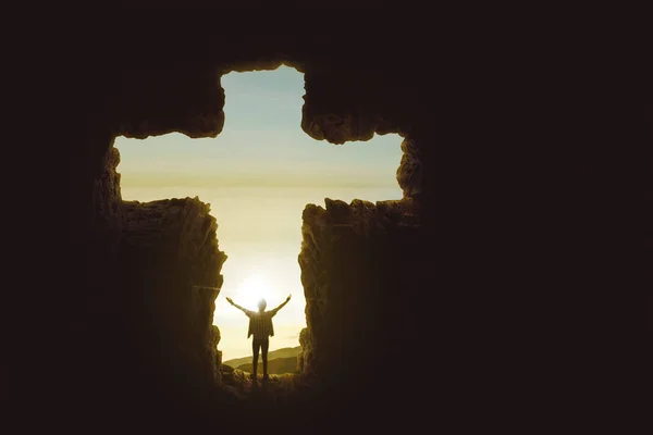 日の出時に洞窟の形をした十字の記号の中に立っている間に手を上げる若い男の後ろのビュー — ストック写真