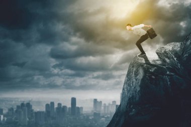 Bulutlu arka planda kayalıklardan atlarken bir bavul taşıyan işadamının yan görüntüsü