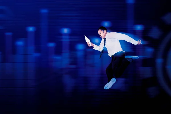 Excited Asian male manager in smart casual clothes jumping and looking at tablet screen against futuristic background