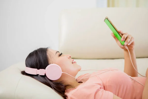 身着现代无线耳机的和平女孩坐在舒适的沙发上 一边听音乐一边看智能手机 — 图库照片