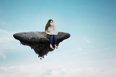 Asyalı iş kadını uçan adada oturur ve mavi gökyüzünde dizüstü bilgisayar kullanır. Yeni iş kurma kavramı