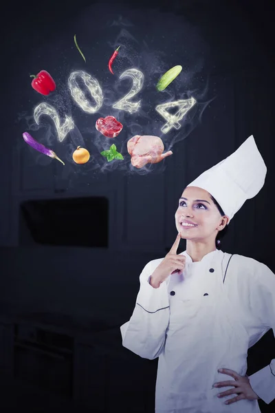 Portrait of a female magician chef with flying food ingredients and smoke shaped of 2024 new year numbers in the kitchen