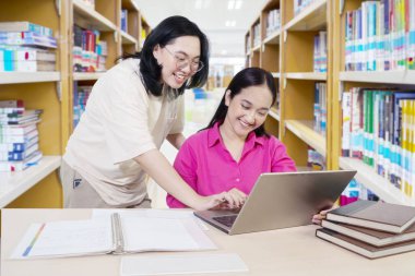 Dizüstü bilgisayara bakan, iletişim kuran gülümseyen gençler, masadaki iki Asyalı kız öğrenci kütüphane arka planında dizüstü bilgisayarlarıyla çalışıyorlar.