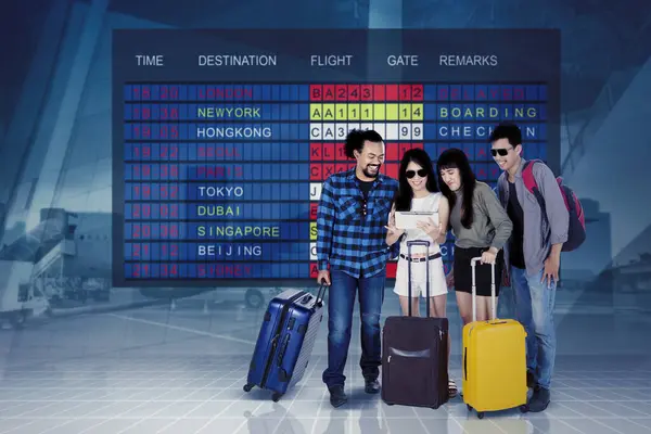 バックグラウンドで飛行スケジュール上のデジタルタブレットを見ているスーツケースを持つ若い旅行者のグループ — ストック写真