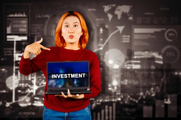 金融チャートの背景の上のスクリーン上の投資アイコンとラップトップコンピュータを示すショックを受けたアジアの女性 — ストック写真
