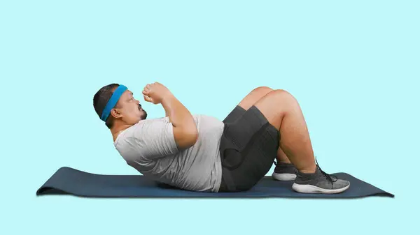 在工作室里做仰卧起坐时穿着运动服的肥胖男人的侧视图 与蓝色背景隔离 — 图库照片