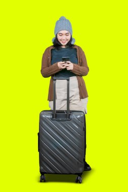 Akıllı telefon kullanan ve beyaz arka planda izole edilmiş bavulu tutan neşeli genç Asyalı kadın.