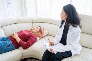 Psikoterapist, Asyalı genç kadını dikkatle dinliyor. Kanepeye uzanıyor ve panoya yazıyor.