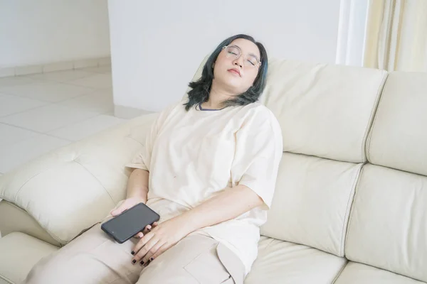 Exhausted Asian millennial girl fell asleep in her sofa while holding a mobile phone