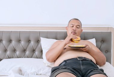 Çizburger ve patates kızartması yerken yatakta yatan kilolu adam.