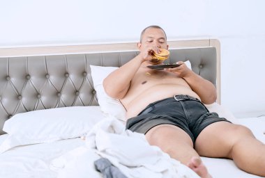 Çizburger ve patates kızartması yerken yatakta dinlenen kilolu adam.