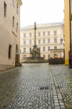 A moody autumn view of the square in Cesky Krumlov, featuring the Baroque Plague Column with the statue of Virgin Mary Immaculata, surrounded by mist, rain, and a tranquil atmosphere clipart