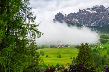 İtalya 'nın Dolomiti (Dolomiti, Dolomiten) kentindeki manzaralı dağ köyü. Yeşil yamaçlardaki sıcacık dağlık evler, yukarıdaki yüksek tepeler ve aşağıdaki vadiden geçen bulutlar.