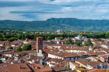 Yukarıdan Lucca, İtalya 'nın panoramik manzarası, kırmızı tuğlalı çatılar ve tarihi kiliseler görünüyor. Toskana tepeleri ve dağları berrak mavi gökyüzünün altında çarpıcı bir yaz ortamı oluşturuyor..
