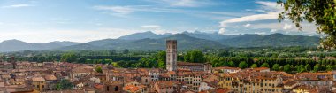 Yukarıdan Lucca, İtalya 'nın panoramik manzarası, kırmızı tuğlalı çatılar ve tarihi kiliseler görünüyor. Toskana tepeleri ve dağları berrak mavi gökyüzünün altında çarpıcı bir yaz ortamı oluşturuyor..