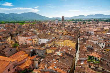 Yukarıdan Lucca, İtalya 'nın panoramik manzarası, kırmızı tuğlalı çatılar ve tarihi kiliseler görünüyor. Toskana tepeleri ve dağları berrak mavi gökyüzünün altında çarpıcı bir yaz ortamı oluşturuyor..