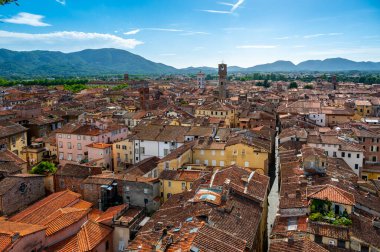 Yukarıdan Lucca, İtalya 'nın panoramik manzarası, kırmızı tuğlalı çatılar ve tarihi kiliseler görünüyor. Toskana tepeleri ve dağları berrak mavi gökyüzünün altında çarpıcı bir yaz ortamı oluşturuyor..