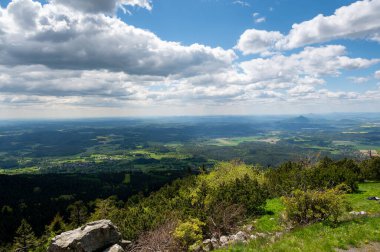 Yeşil tarlaların panoramik manzarası ve Jested yakınlarındaki orman tepeleri, Ralsko dağının keskin zirvesi mavi gökyüzünün altında dağınık bulutlarla görülebilir..