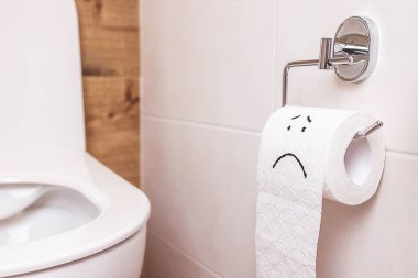 Banyo ve klozetin arka planında kopyalama alanı var. Beyaz duvarda üzgün bir yüz asılı beyaz tuvalet kağıdı rulosu var. Hastalık konsepti, mide sorunları, bağırsaklar, basur.