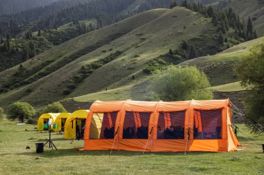 Büyük turuncu bir kamp çadırı manzaralı bir dağ vadisinde kurulmuş, güneş ışığıyla yıkanmış, doğayla çevrili.