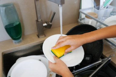 Eller, mutfak lavabosunun altında akan süngerle beyaz tabak temizler. Ev işleri kavramı..