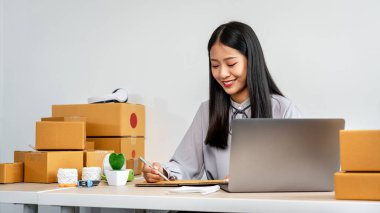 Genç kadın küçük işletme girişimcisi evde çalışıyor çevrimiçi müşteriden sipariş veriyor ve müşteriye gönderilen ürünleri paketlerken verileri kontrol ediyor.