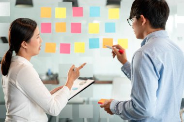 İki işletme yöneticisi, ofiste birlikte pazarlama planının planlanmasına yönelik yeni bir iş projesi hakkında fikir alışverişinde bulunmak için kilit noktayı duvara yazıp not alıyorlar..