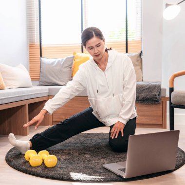 Asyalı bir kadın halterle yerde oturuyor ve evde oturma odasında egzersiz yapmadan önce internetten yoga dersi alırken dizüstü bilgisayar kullanıyor.
