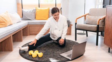 Asyalı bir kadın halterle yerde oturuyor ve evde oturma odasında egzersiz yapmadan önce internetten yoga dersi alırken dizüstü bilgisayar kullanıyor.