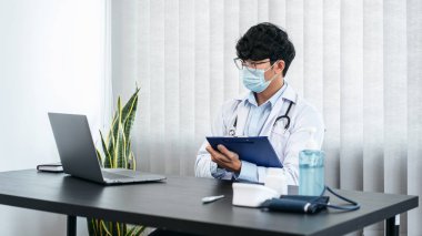 Yüz maskesi takan erkek doktor, tedavi hakkında bilgi aramak için dizüstü bilgisayar kullanıyor. Hastanın semptomları ve klinikte çalışırken reçete yazmak için pano tutuyor.