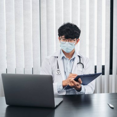 Yüz maskesi takan erkek doktor, tedavi hakkında bilgi aramak için dizüstü bilgisayar kullanıyor. Hastanın semptomları ve klinikte çalışırken reçete yazmak için pano tutuyor.