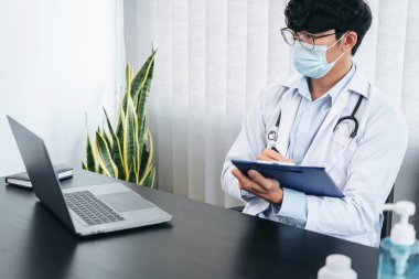 Yüz maskesi takan erkek doktor, tedavi hakkında bilgi aramak için dizüstü bilgisayar kullanıyor. Hastanın semptomları ve klinikte çalışırken reçete yazmak için pano tutuyor.