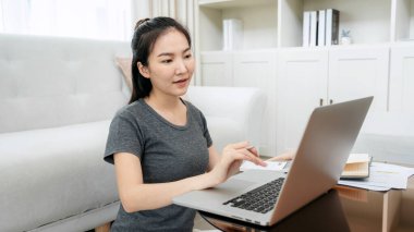 Müşteriden sipariş almak için dizüstü bilgisayar kullanan Asyalı serbest kadın ve çevrimiçi pazarlama teslimatı sırasında ürünleri kontrol ederken ev ofisteki defterde müşteri bilgileri yazıyor.
