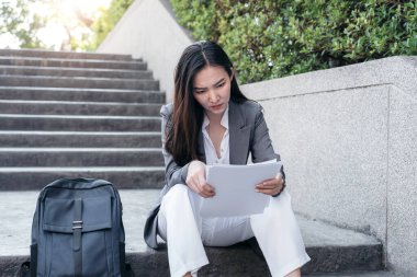 Takım elbiseli Asyalı iş kadını, elinde iş raporu belgesi tutarken ve mali tabloları okurken ofis dışındaki merdivenlerde yeni başlatılan projenin analizini yaparken.  