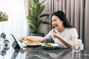 Sağlıklı yemek için sebze salatası yiyen Asyalı kadın tablete dokunurken arkadaşlarıyla sohbet ederken ve evde yemek odasında film izlerken.