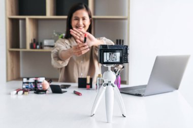 Mutlu gülümseyen zarif kadın blogcu, makyaj malzemesi kozmetik inceleme ürünü sunuyor ve kamera ekranında sosyal ağa canlı yayın yapan video bloglarını öğretiyor..