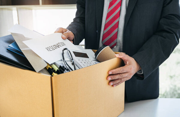 Businessman carrying packing up all his personal belongings and files into a brown cardboard box to resignation in modern office, resign concept.