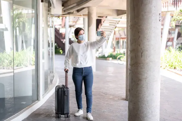 Tourist woman with luggage using smartphone to taking selfie photo travelling and wearing medical face mask to protection from coronavirus and travel with new normal lifestyle during covid-19 outbreak