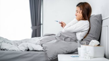 Asyalı bir kadın yüzüne dokunmak için elini kullanırken ve battaniyede otururken vücut ısısını ölçmek için termometreyi kullanırken ilaç kullanırken kendini hasta ve baş ağrısı hissediyordu.