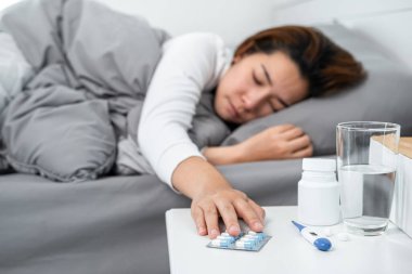 Asyalı kadın kendini hasta hissediyor ve vücut ısısını ölçmek ve ilaç almak için termometre kullandıktan sonra yatakta battaniyede yatarken masadaki ilaçlara dokunmak için ellerini kullanıyor.