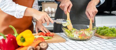 Asyalı romantik çift salatalık ve domatesi ahşap kesim tahtasına koyarken genç adam kepçeyle evde sağlıklı yemek pişirmek için sebze salatası hazırlıyor.