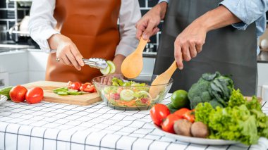 Asyalı romantik çift salatalık ve domatesi ahşap kesim tahtasına koyarken genç adam kepçeyle evde sağlıklı yemek pişirmek için sebze salatası hazırlıyor.