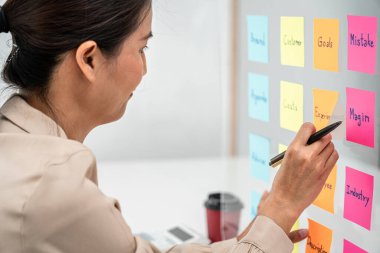 İş kadını elinde kalem tutarken ve ofisteki cam bir duvarda fikir paylaşımı yaparken, iki iş adamı yeni bir başlangıç projesiyle birlikte pazarlama planını planlamaya çalışıyor.