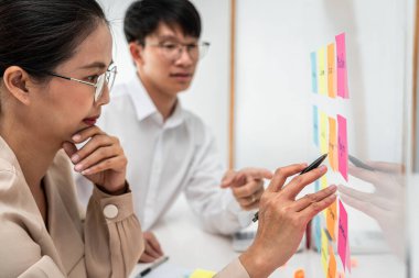 İş kadını elinde kalem tutarken ve ofisteki cam bir duvarda fikir paylaşımı yaparken, iki iş adamı yeni bir başlangıç projesiyle birlikte pazarlama planını planlamaya çalışıyor.